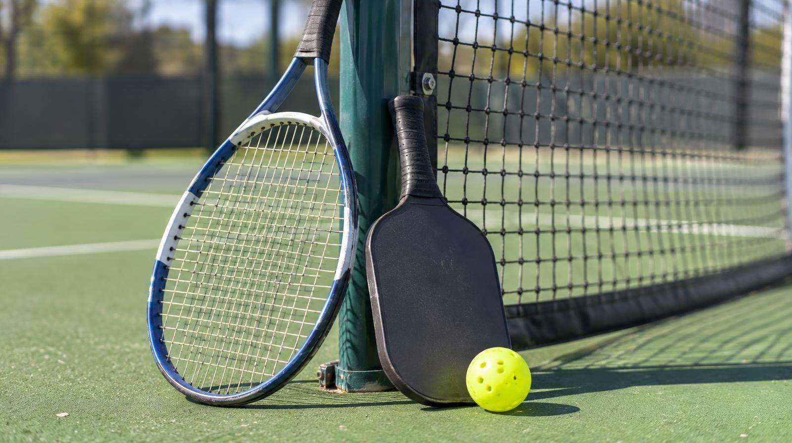 Tennis racket and pickleball paddle resting on a green court near a net post.