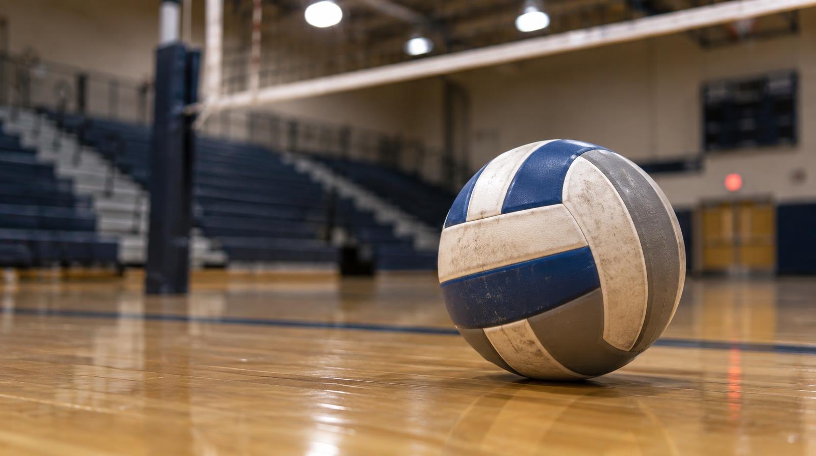 A volleyball sitting on a hardwood gym floor near the net, bleachers blurred behind.