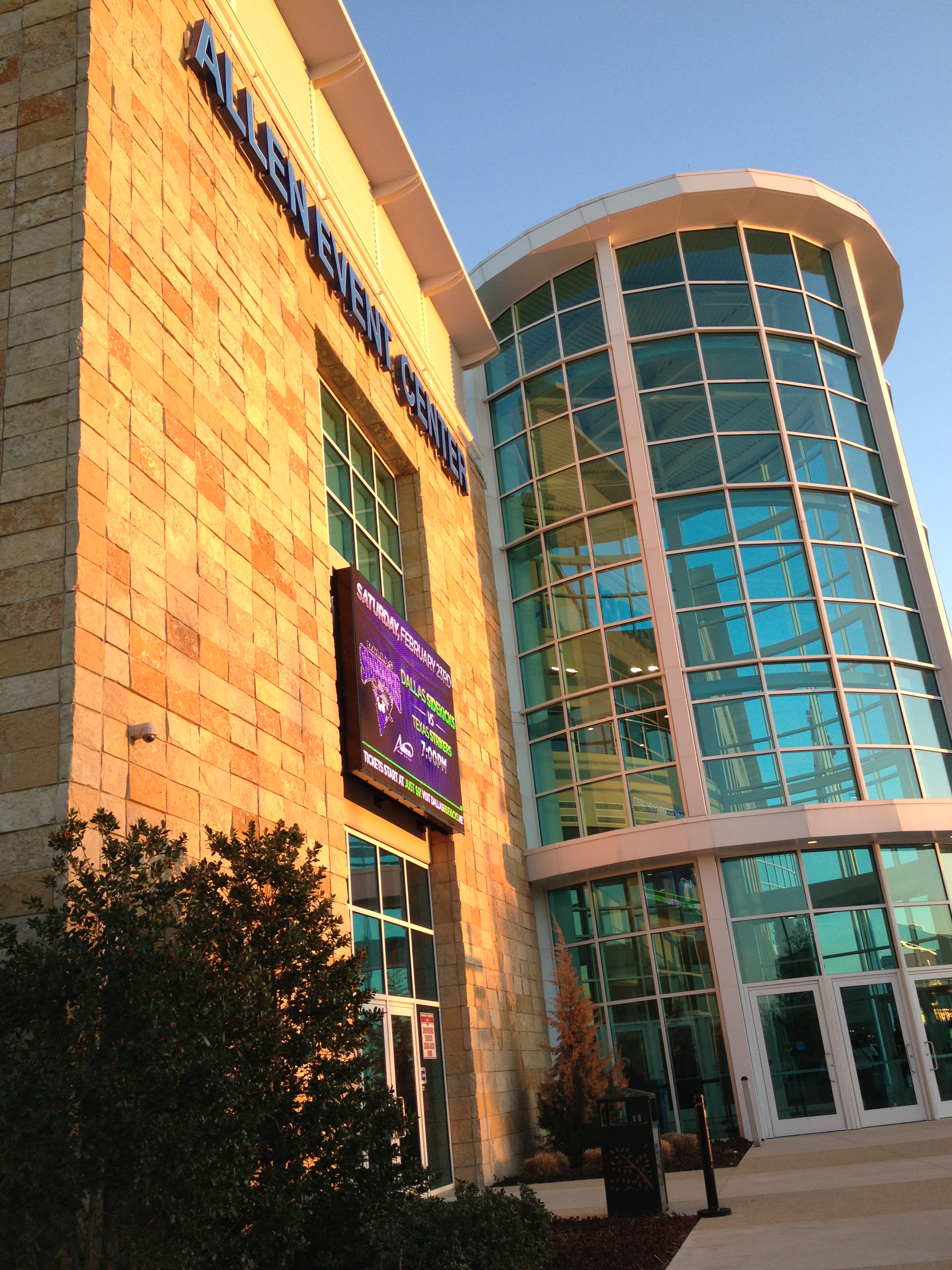 Exterior of an arena with a tan stone facade, digital marquee, and curved glass entrance.
