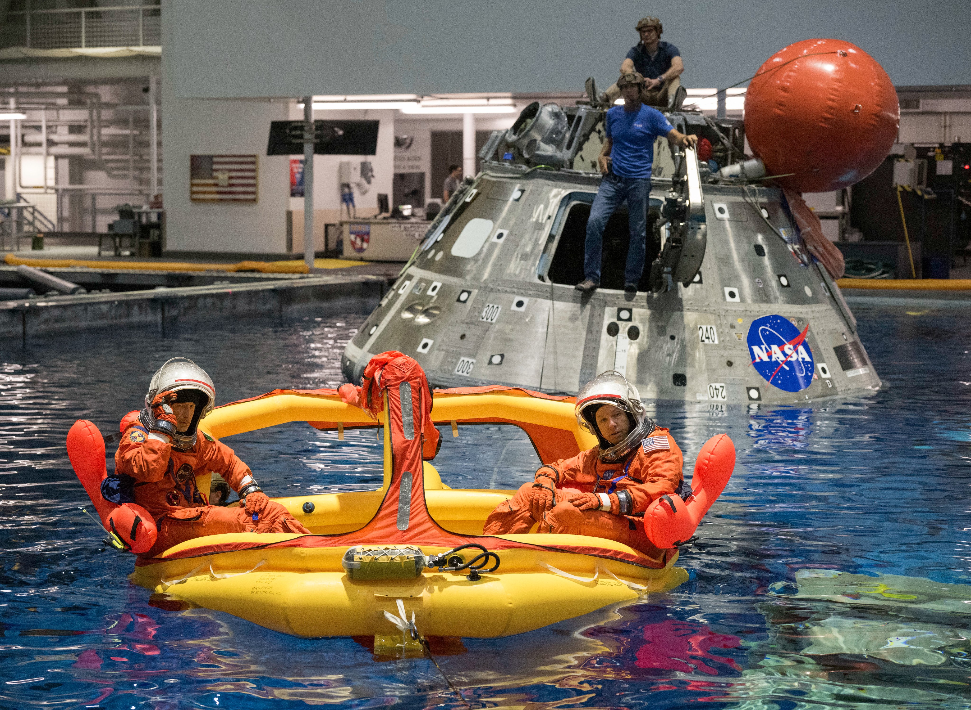 Two astronauts in orange spacesuits sit in an inflatable raft during a water training exercise, with a spacecraft capsule and NASA equipment in the background.