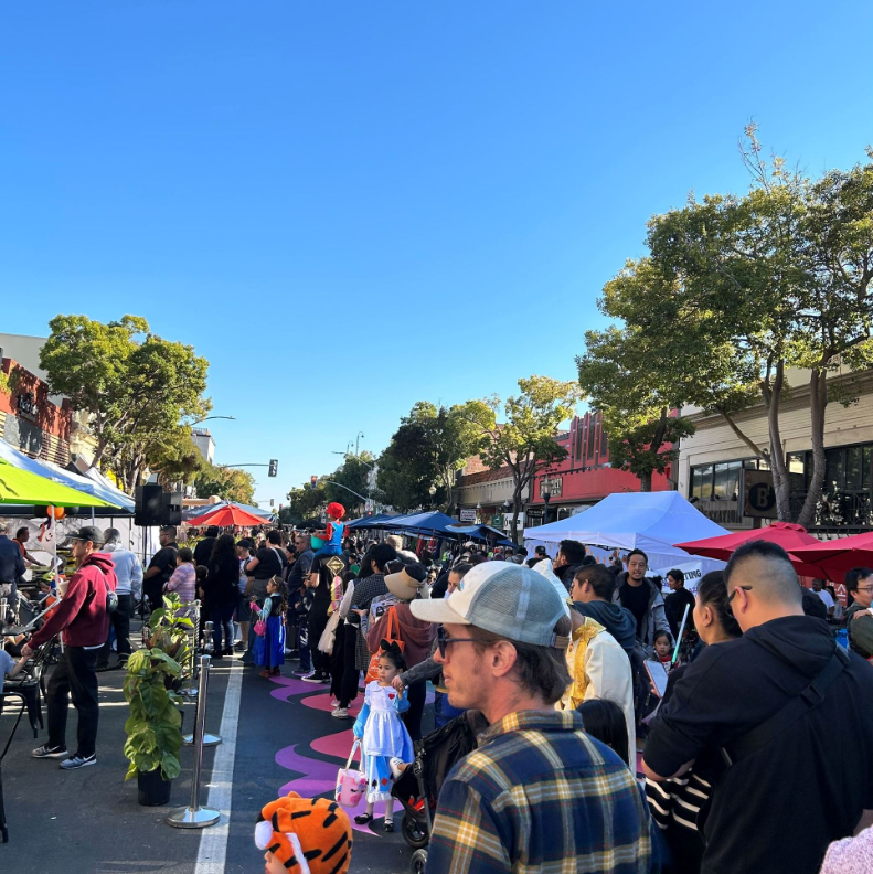 Families and children in costumes crowd B Street beneath colorful vendor tents.