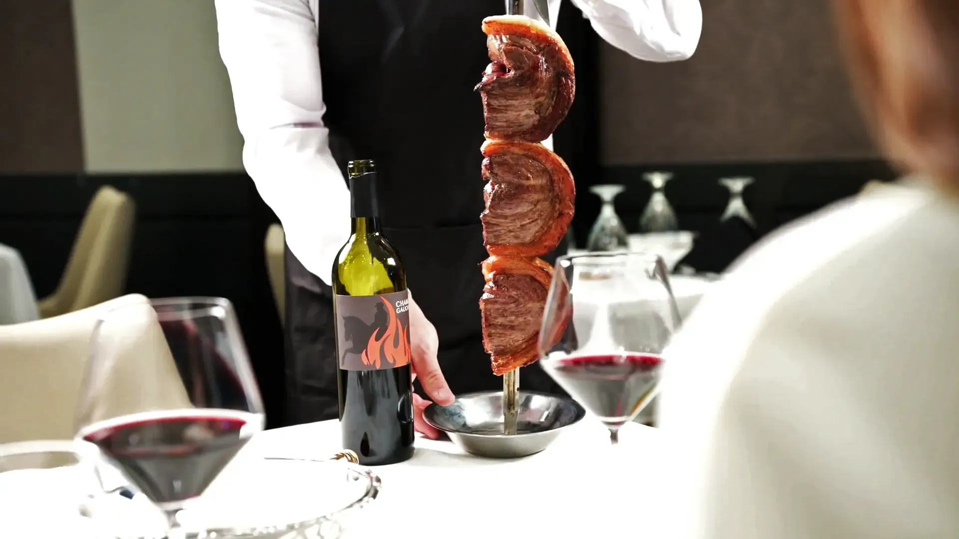 A server presents a skewer of grilled meat tableside next to a wine bottle at an elegant restaurant table set with glasses of red wine.