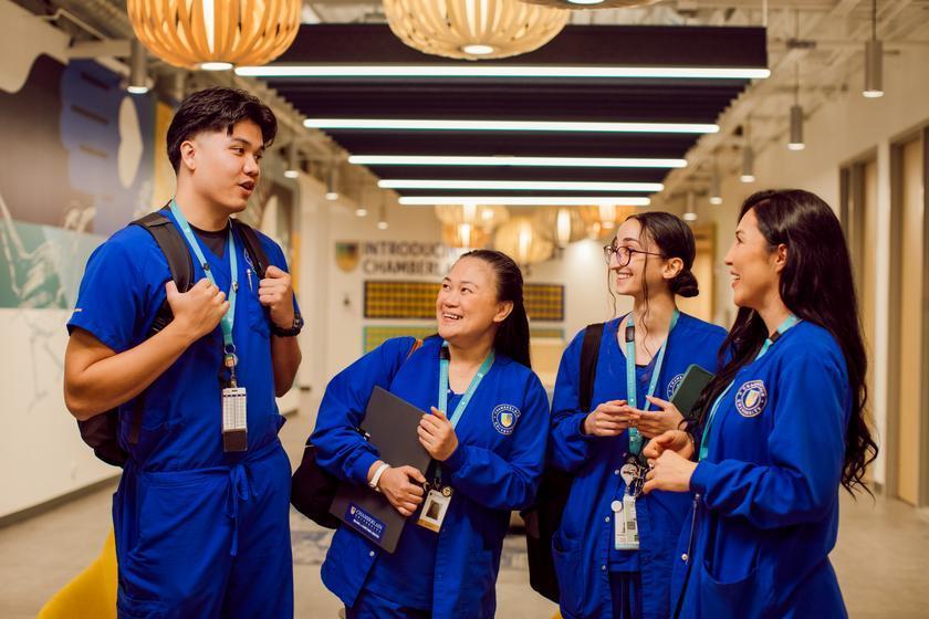 Four nursing students in blue uniforms and backpacks talk and smile together in a school hallway.