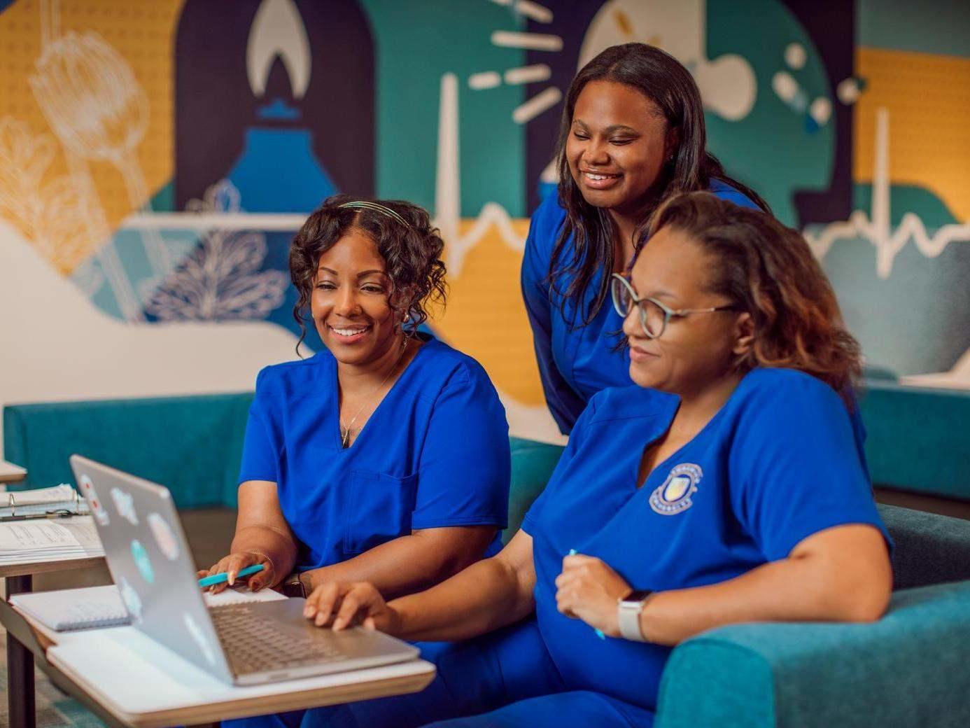 Three women in blue medical scrubs gather in a colorful lounge, with two seated at a laptop and another standing behind them.