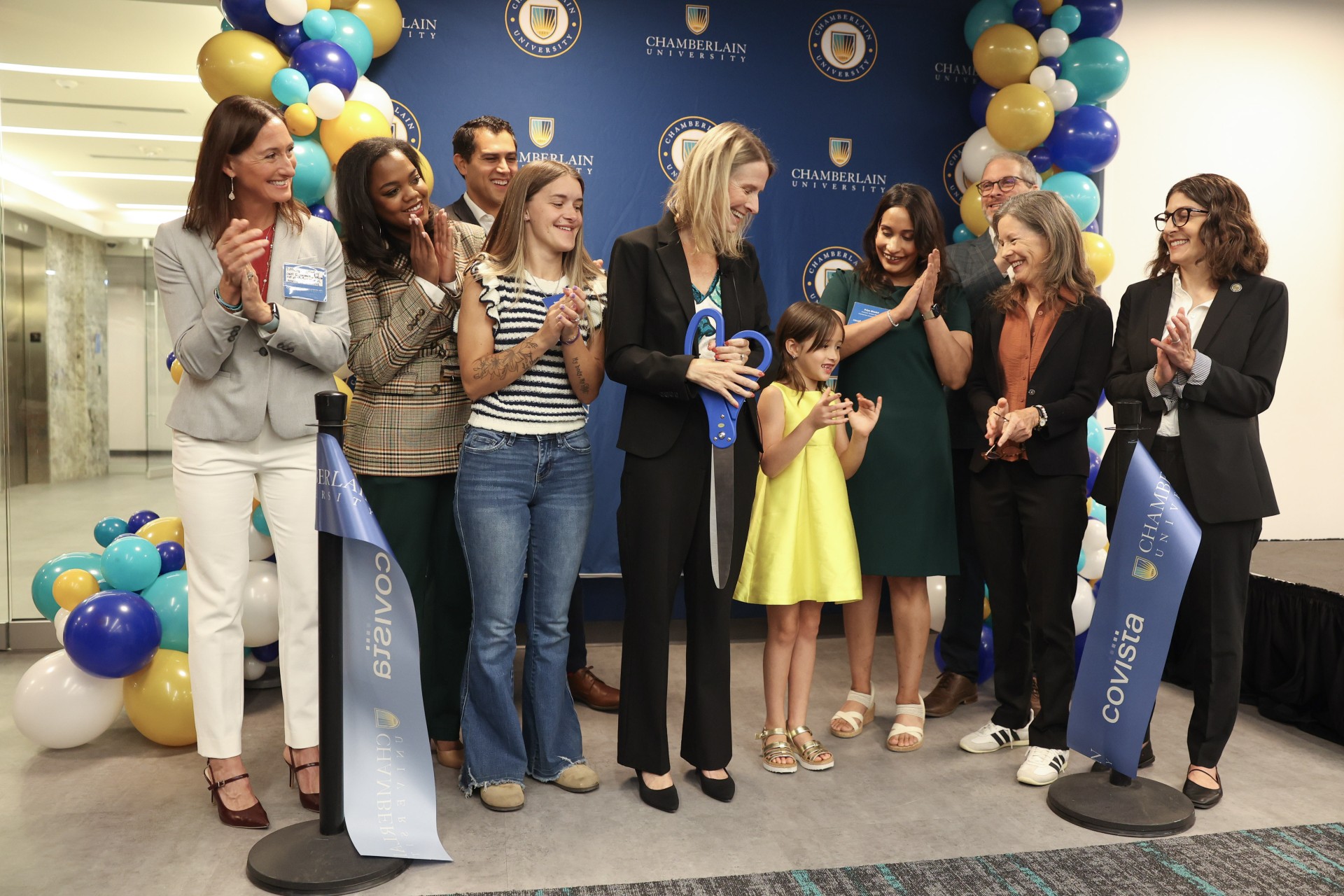 A group of people pose together as a woman cuts a ribbon with oversized scissors at a Chamberlain University and Covista ribbon-cutting ceremony, with balloon decorations in blue, gold, and white in the background.