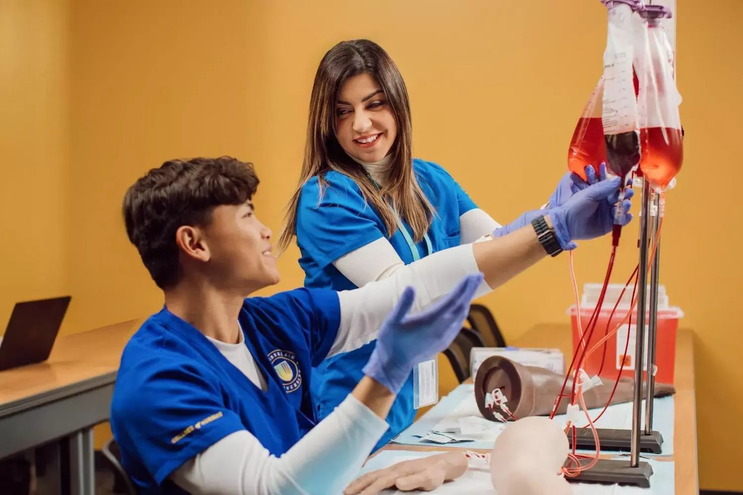 Two nursing students in blue scrubs and gloves practice handling an IV blood bag and tubing in a clinical training setting.