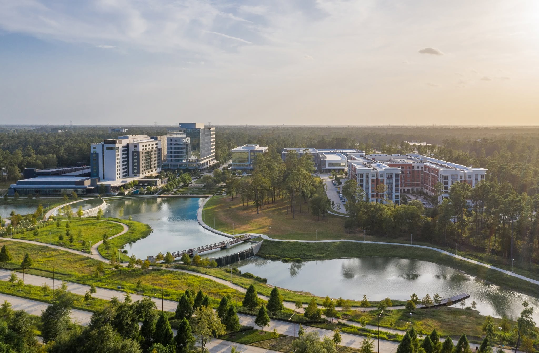 Aerial view of a modern mixed-use development with office buildings, apartments, and a landscaped park surrounding a curving lake, set against a backdrop of dense forest.