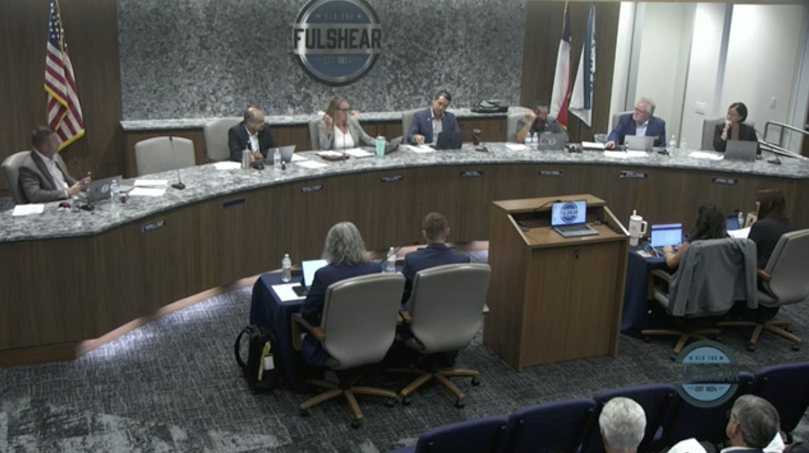 Photo inside council meeting shows men and women sitting in gray chairs.,