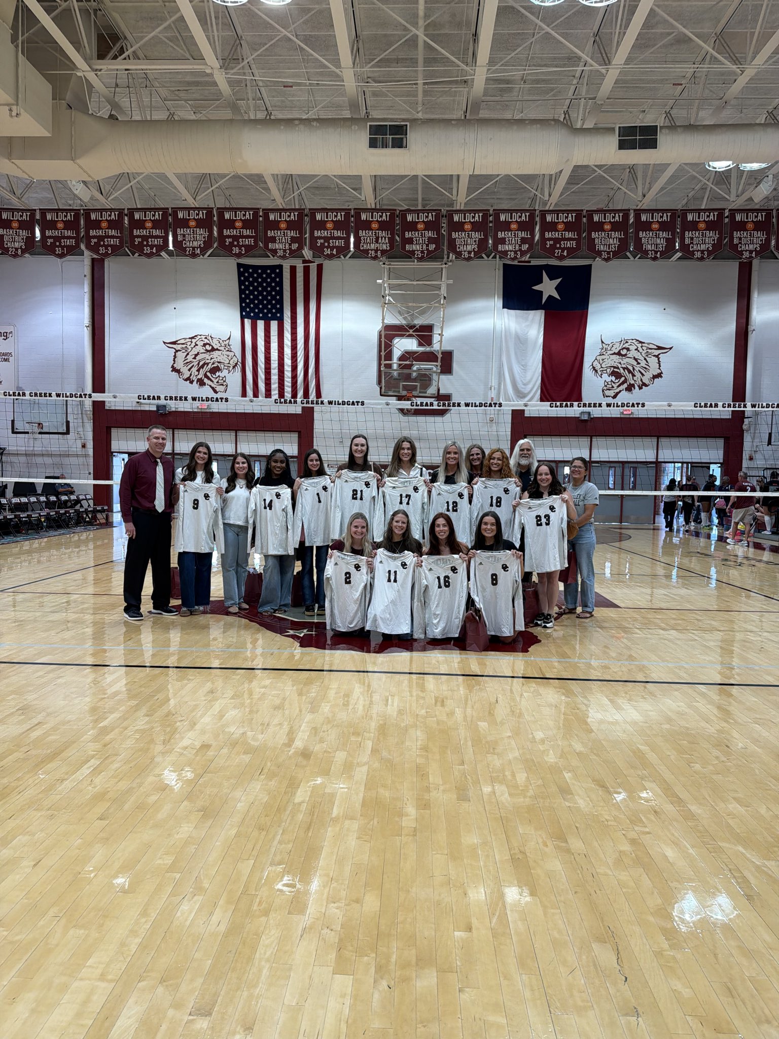 A group of Clear Creek volleyball players and coaches pose on a gym court, holding white jerseys with player numbers, beneath championship banners, American and Texas flags, and Clear Creek Wildcats logos.