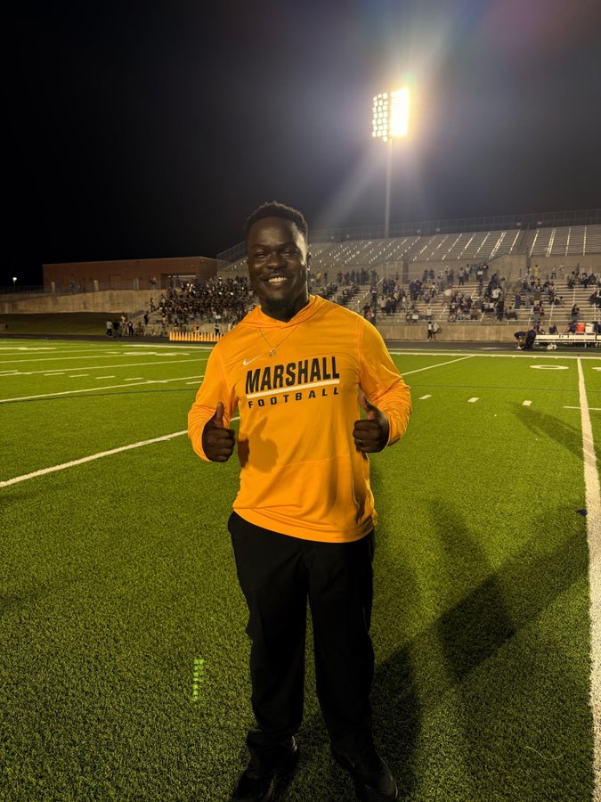 Man in yellow sweatshirt standing on football field with thumbs up.