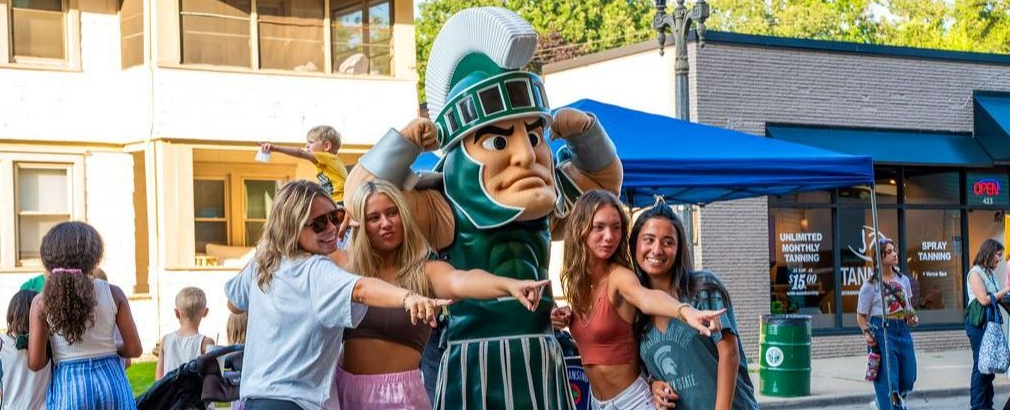 The MSU Spartan mascot, Sparty, poses with a group of children and young women on a downtown street during a past community event.