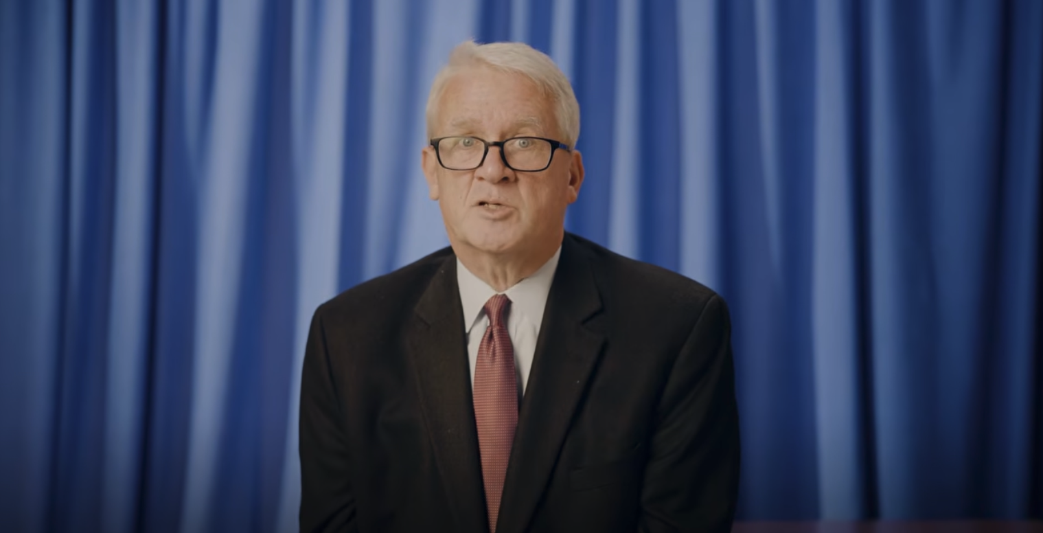 A man with white hair and glasses wears a dark suit and red tie while speaking in front of blue curtains.