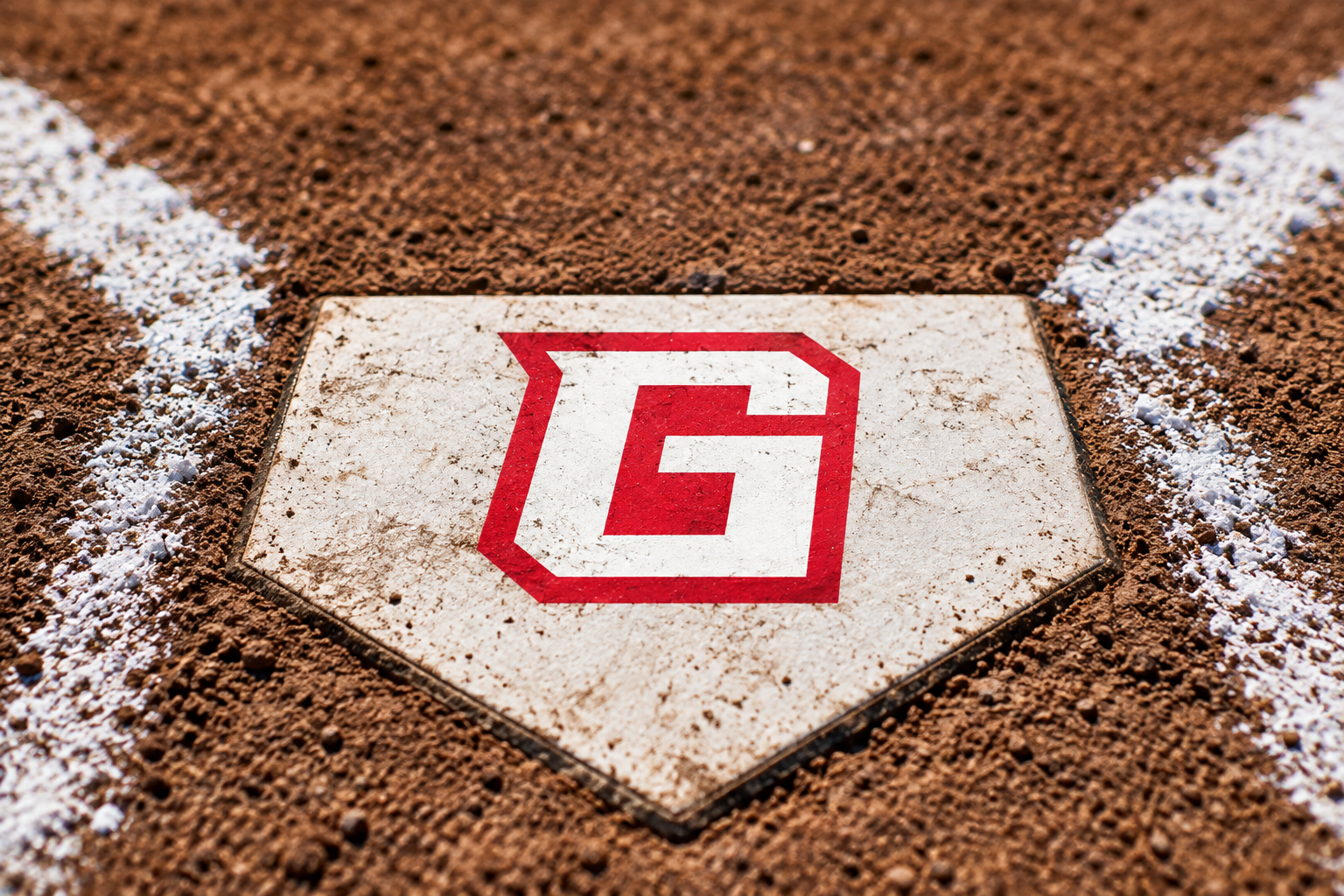 Close-up of a baseball home plate featuring a red and white G logo, surrounded by dirt and chalk baselines on a baseball field.