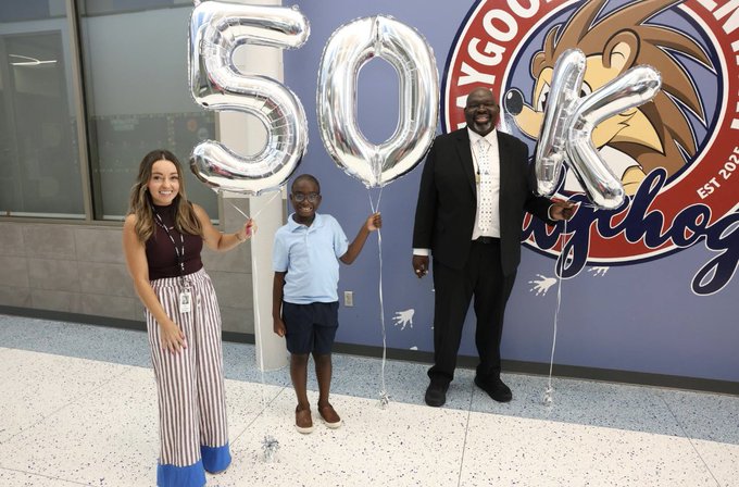 A group of three people stands inside a school near a large school mascot mural. They hold silver balloons spelling “50K,” celebrating a milestone. The group includes an adult woman, a child, and an adult man in a suit.