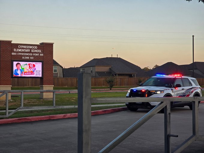 School entrance at dusk with a law enforcement patrol vehicle parked outside and its emergency lights flashing.