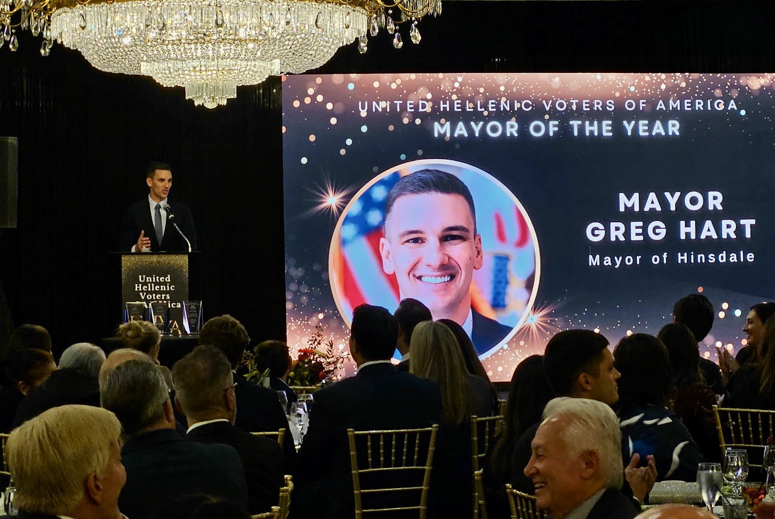 A man speaks at a podium beside a screen honoring Hinsdale Mayor Greg Hart as the United Hellenic Voters of America’s Mayor of the Year.