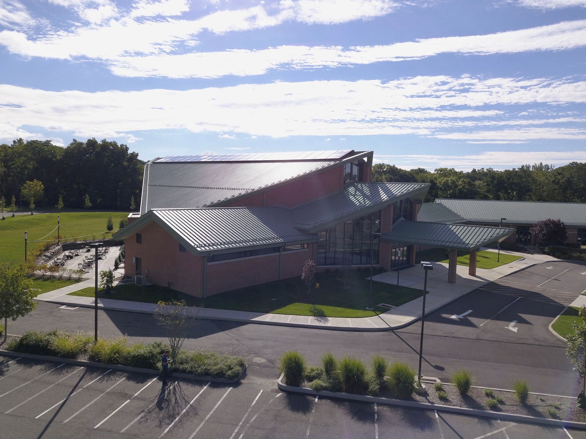 Aerial drone photo of a modern brick building with angular green metal roofing, solar panels, tall glass windows, and a covered entrance, surrounded by a parking lot, grassy lawn, and trees under a partly cloudy sky.
