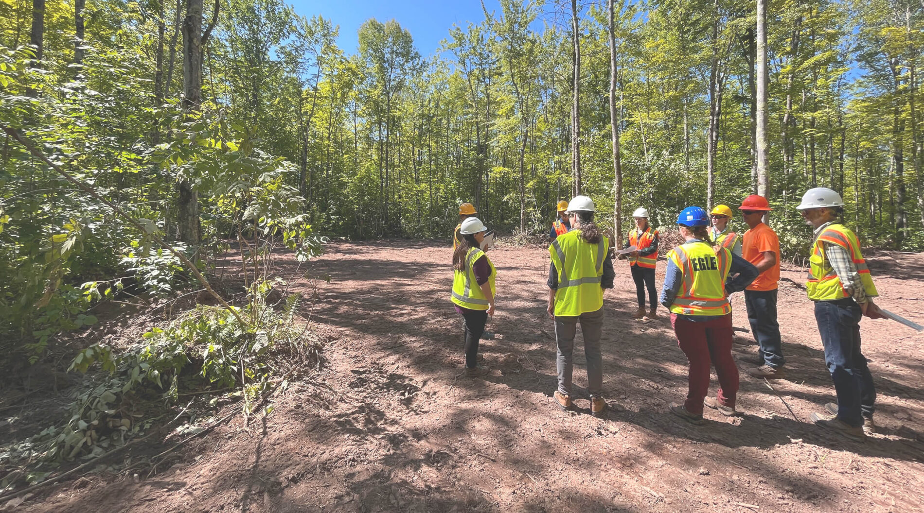 A group wearing hard hats and high-visibility safety gear gathers on a cleared dirt site surrounded by dense woodland.