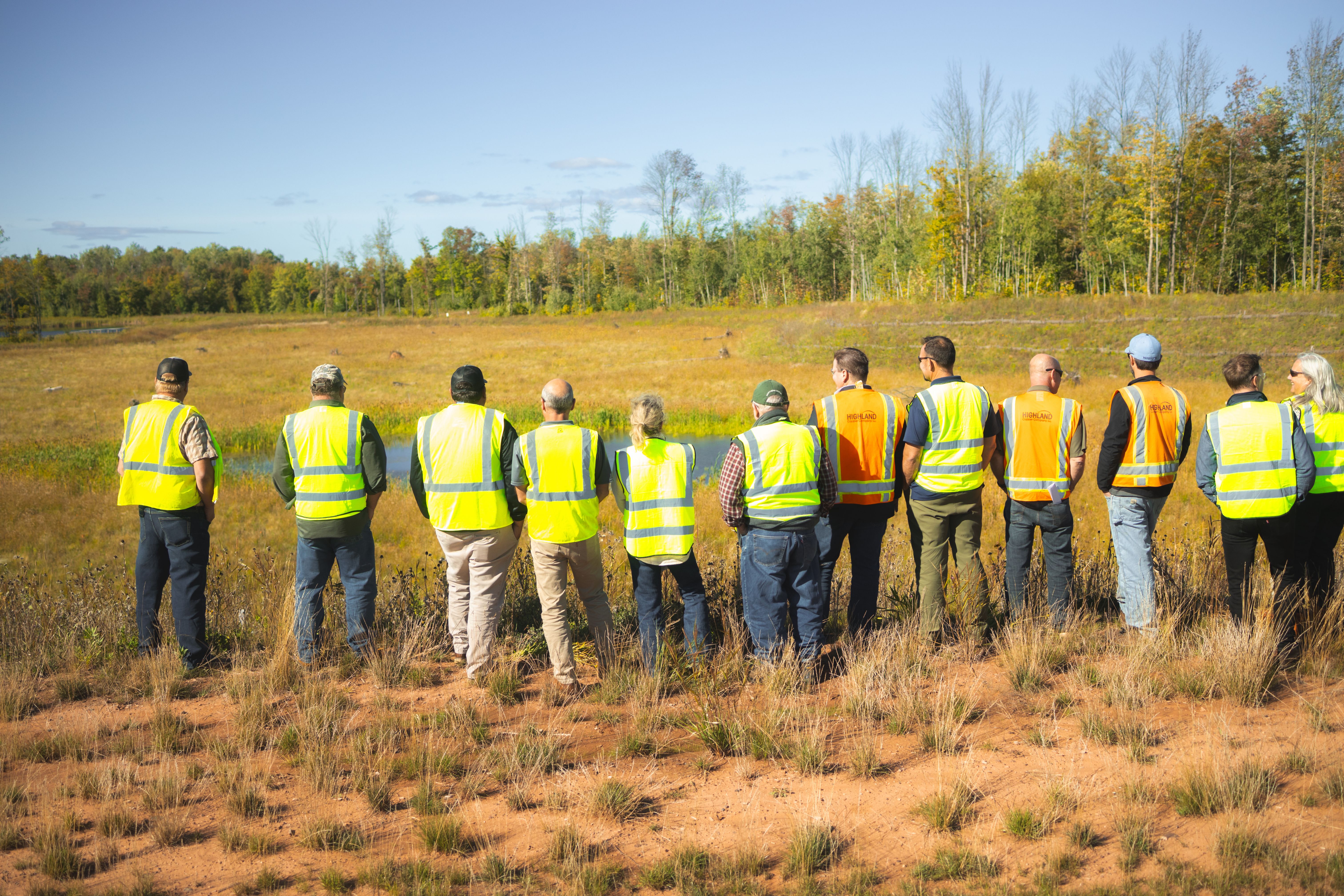 A group of people in high-visibility safety vests stands in a grassy field overlooking a wooded area.
