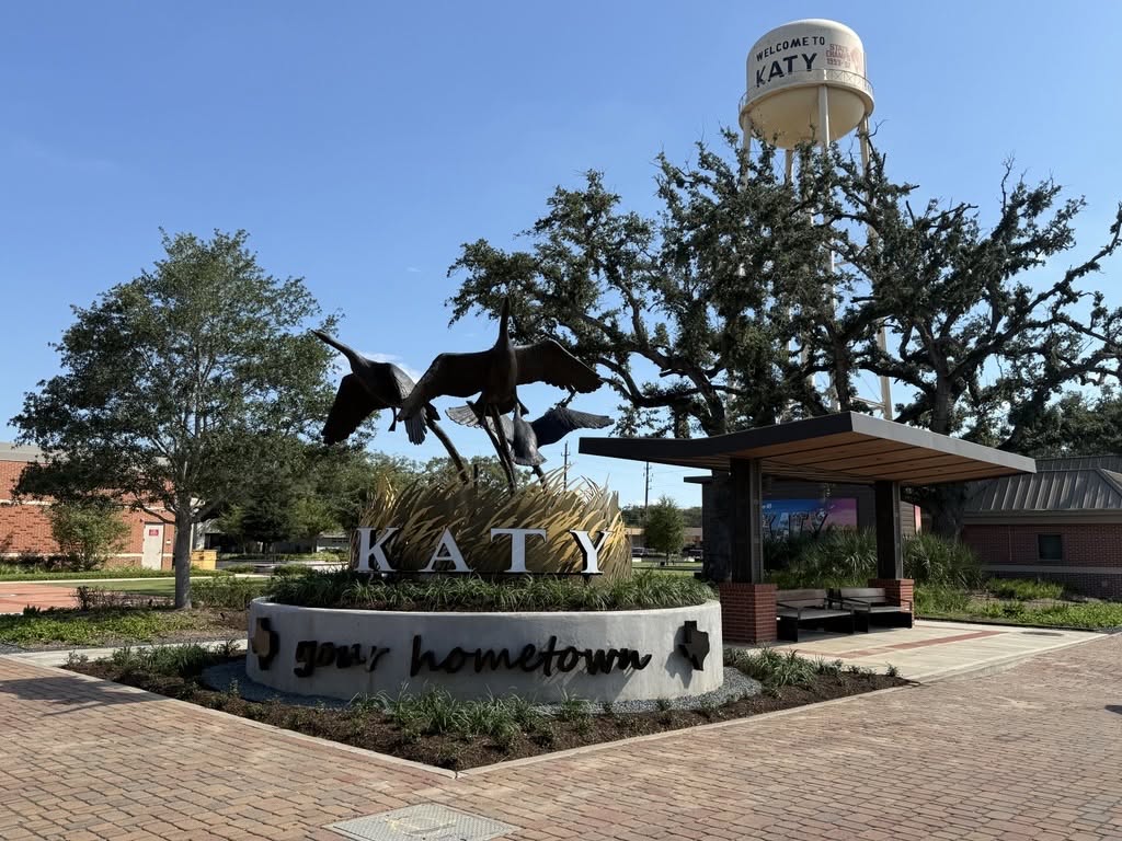 Sculpture of flying geese above a sign reading “Katy your hometown” with a water tower and trees in the background under a clear sky.
