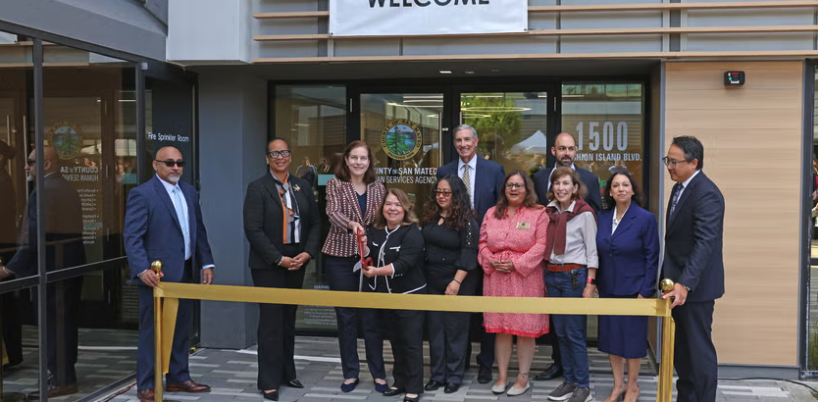 County and city officials and Human Services Agency staff pose for a ribbon-cutting ceremony outside the new Fashion Island office, with a gold ceremonial ribbon in the foreground.