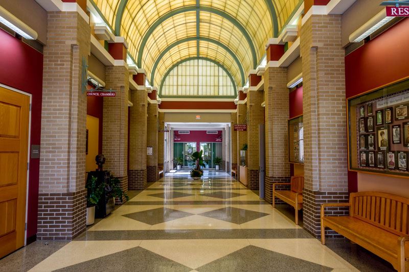 Interior hallway of a municipal building with brick columns, wood benches, geometric tile flooring, and a large arched glass skylight.