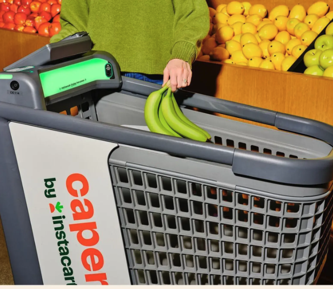 A person places a bunch of bananas into a shopping cart at a grocery store, with bins of apples and lemons visible in the background.