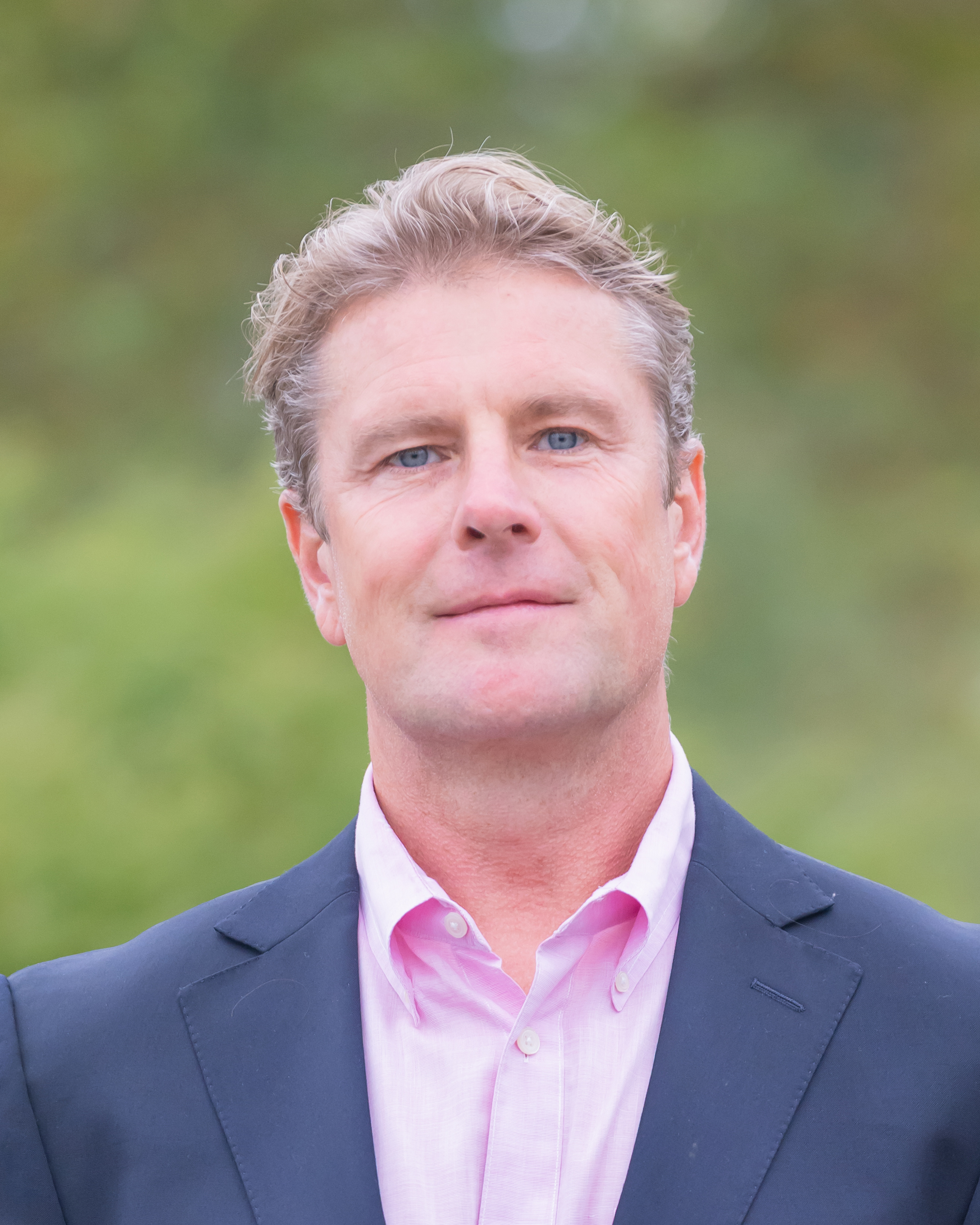 Jeff Frese, wearing a navy blazer and pink button-down shirt, smiles outdoors against a blurred green background.
