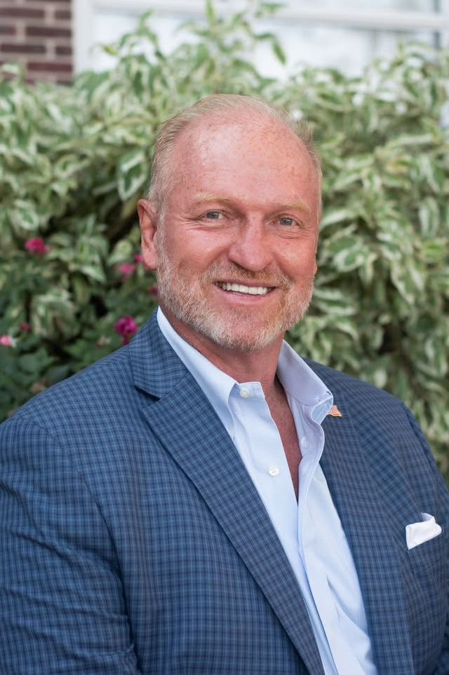 A smiling man wearing a blue checkered blazer and light blue dress shirt, standing in front of greenery.