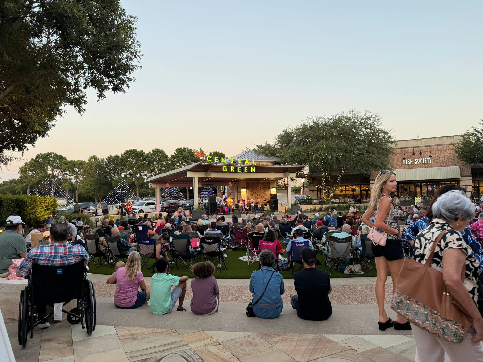 crowd gathers on a lawn at dusk for an outdoor concert at "Central Green," watching a band perform on a stage lit with string lights, with the "Dish Society" restaurant visible in the background.