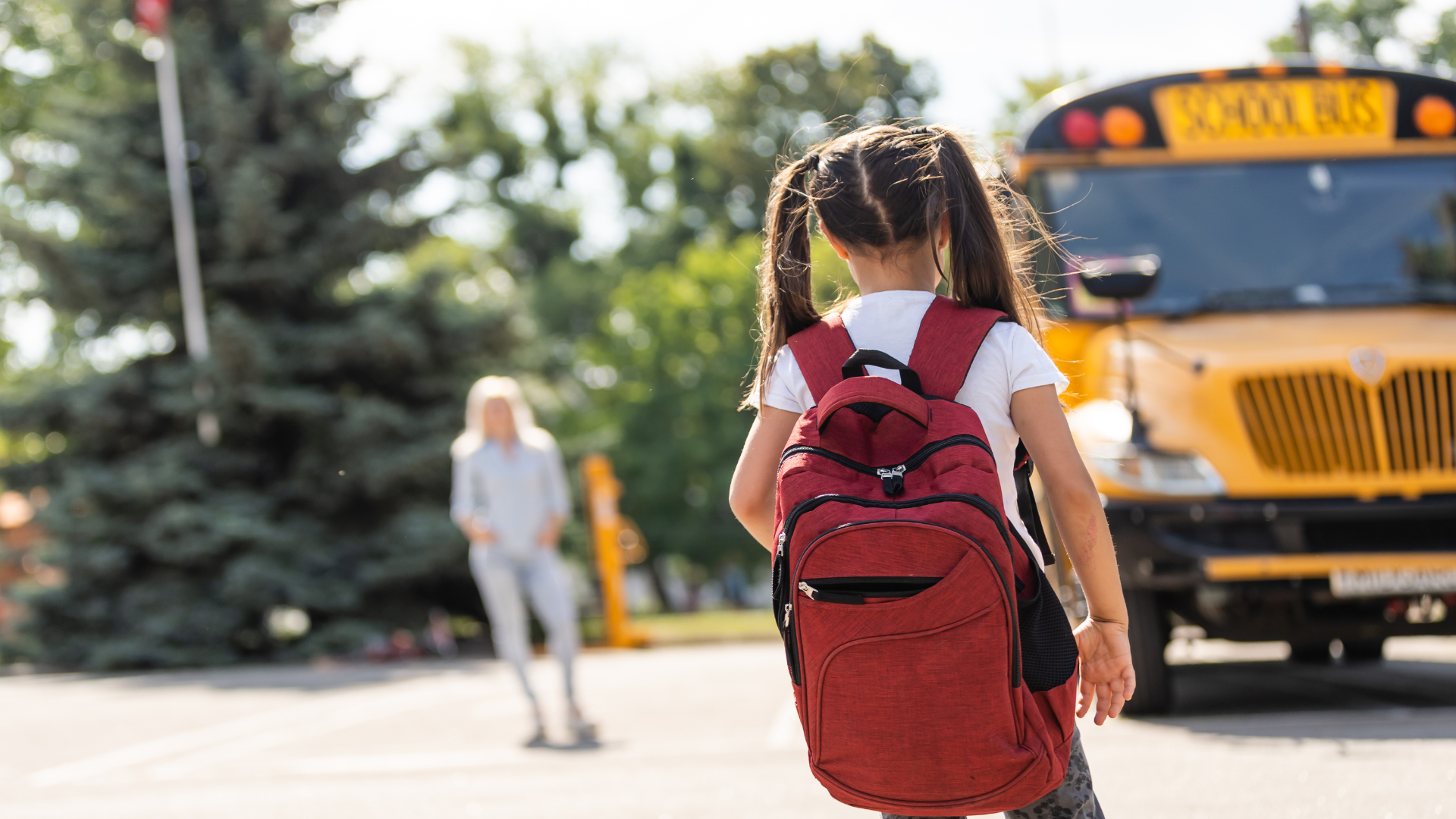 Girl in pigtails walking near school bus towards aide with red backpack on.