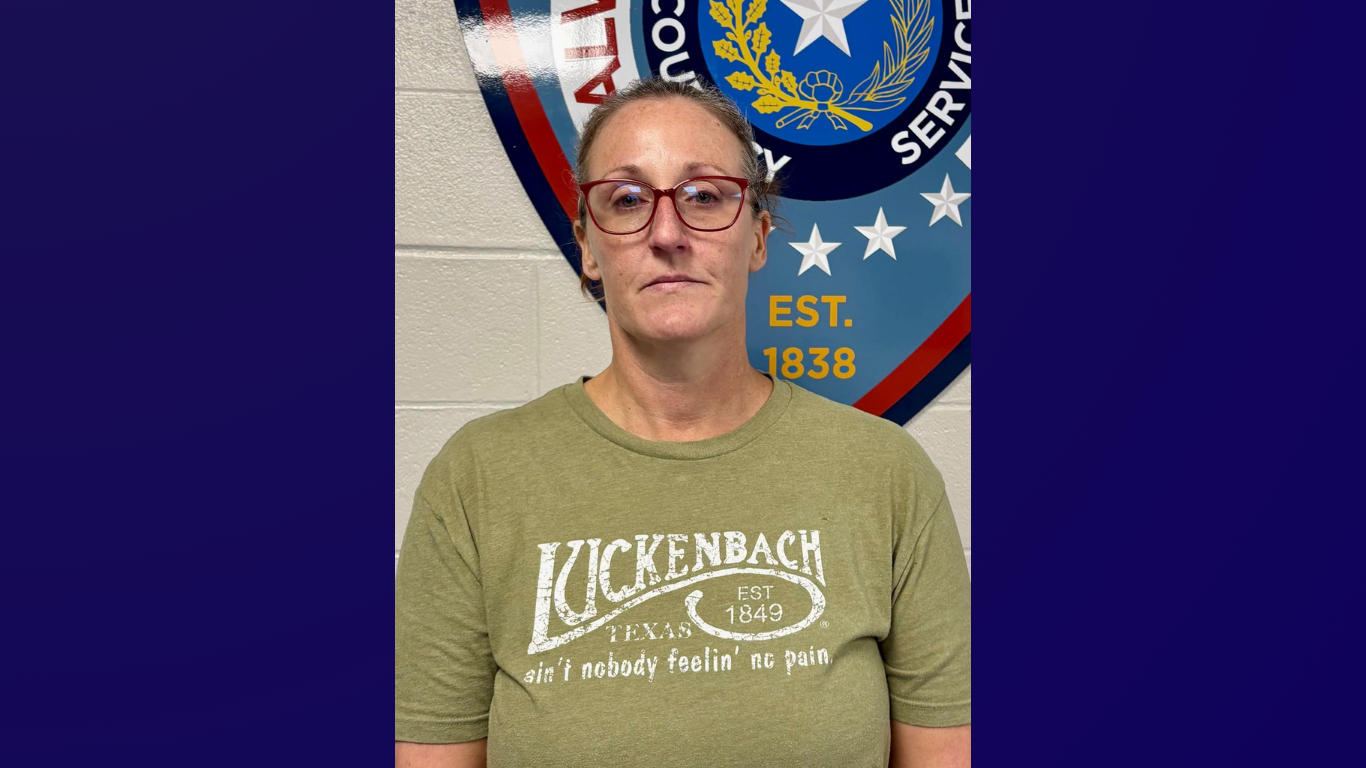 Booking photo of a woman with glasses and hair pulled back, wearing an olive green "Luckenbach, Texas" T-shirt, standing against a wall in front of a county sheriff's office seal reading "Est. 1838."