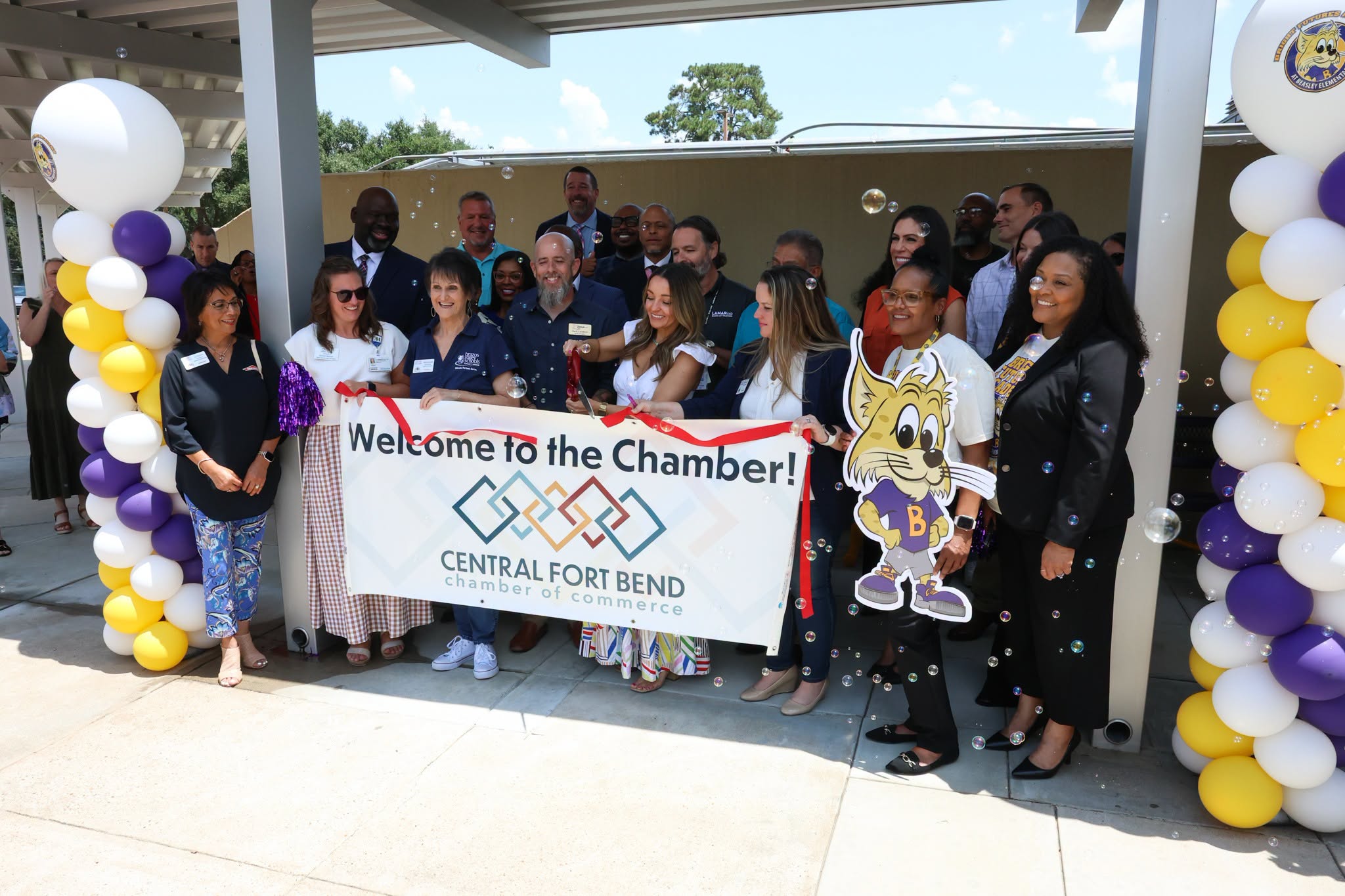 Community members gather at a ribbon-cutting ceremony beneath purple, gold, and white balloon decorations, holding a “Welcome to the Chamber!” banner from the Central Fort Bend Chamber of Commerce.