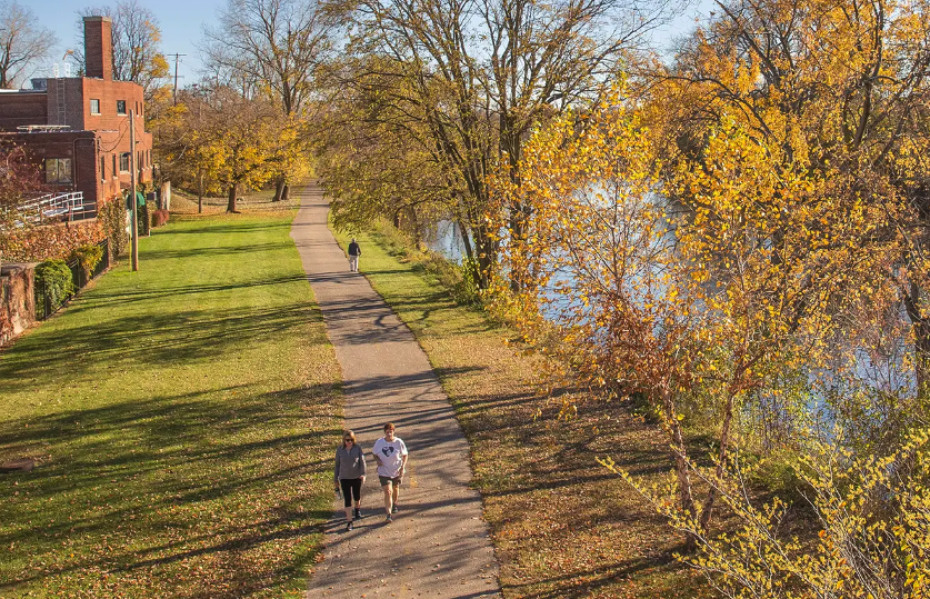 People walk along the Lansing River Trail beside the Grand River, surrounded by trees with fall foliage.