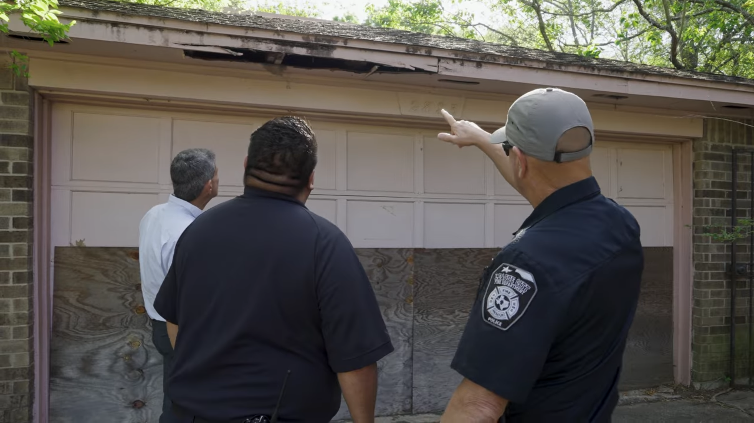 Three officials, including a League City Fire Department member, inspect a deteriorating garage roof and boarded-up door during a code enforcement walkthrough.