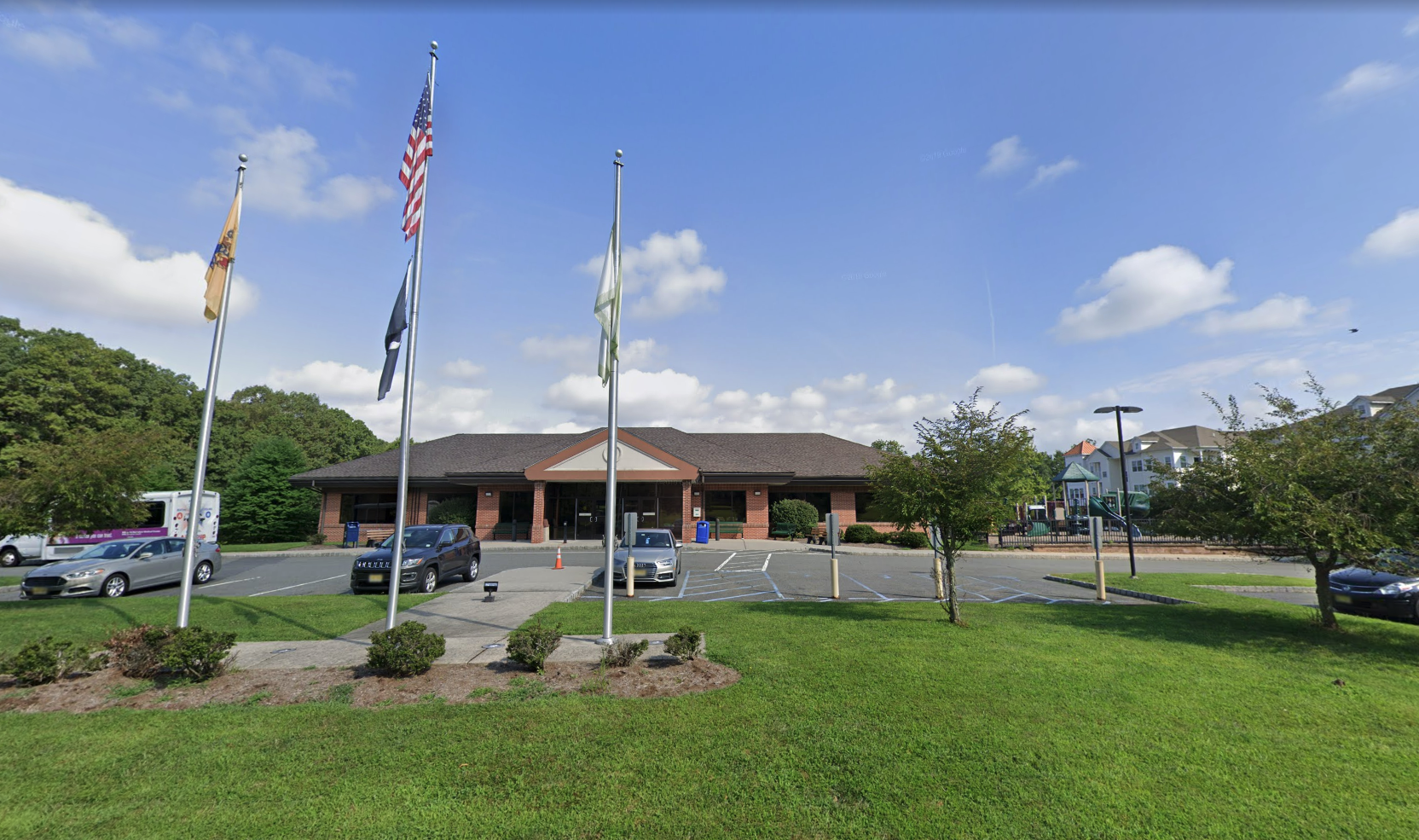 A brick public building with several flagpoles, a parking lot and a playground in the background.
