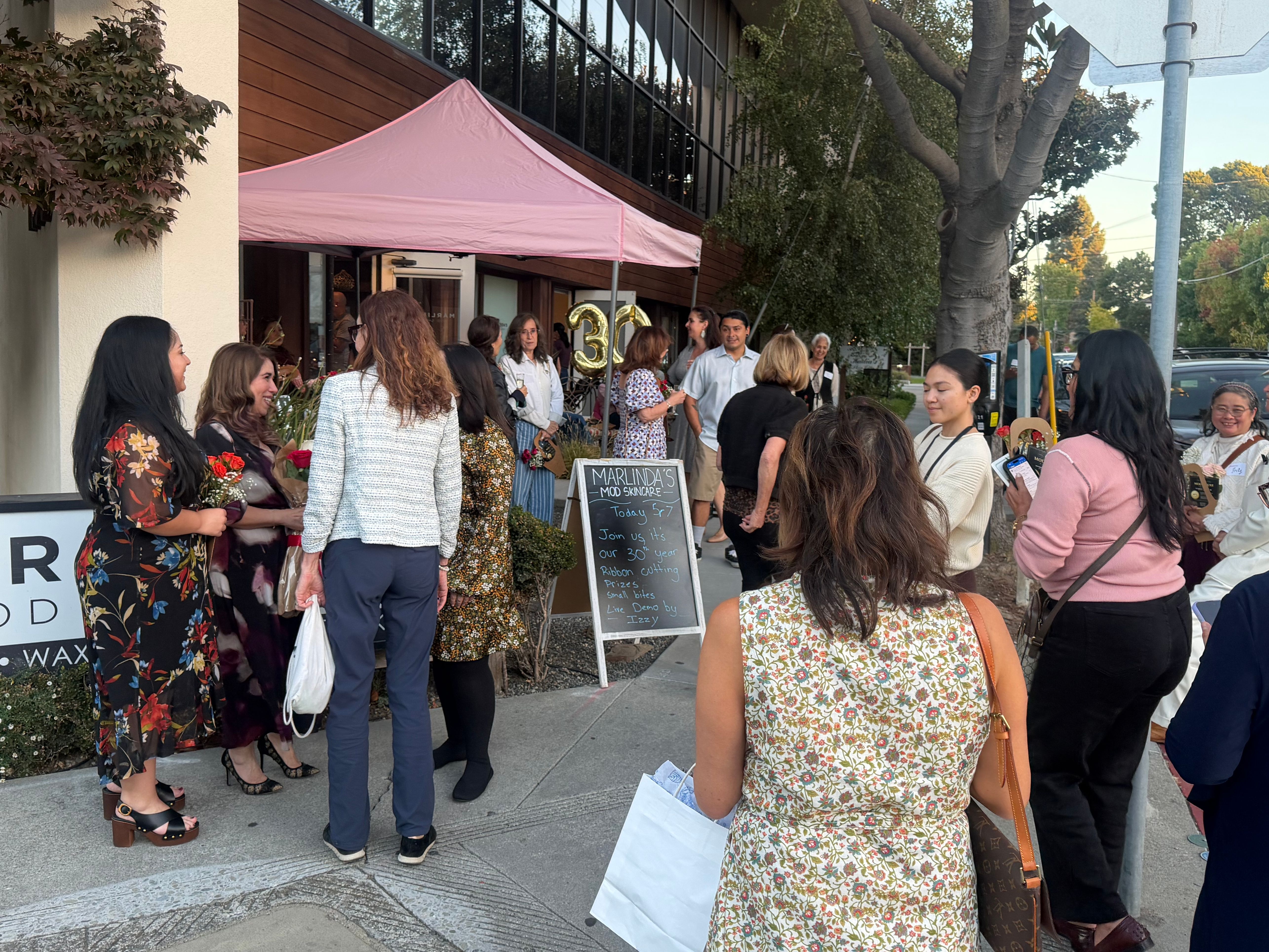 Guests gather beneath a pink canopy outside a spa beside gold anniversary balloons and a chalkboard sign.