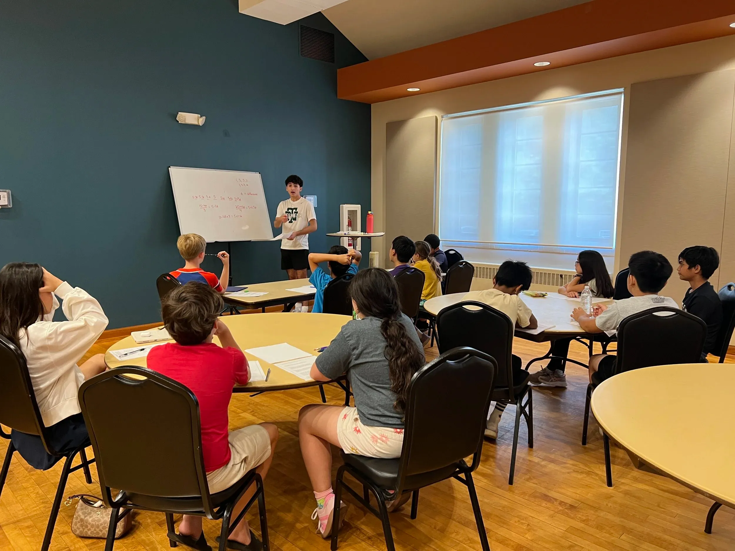 A young person presents beside a whiteboard with math equations while a group of students sits around tables with worksheets.