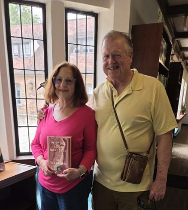 A man and woman stand together indoors near leaded-glass windows in a historic hall.