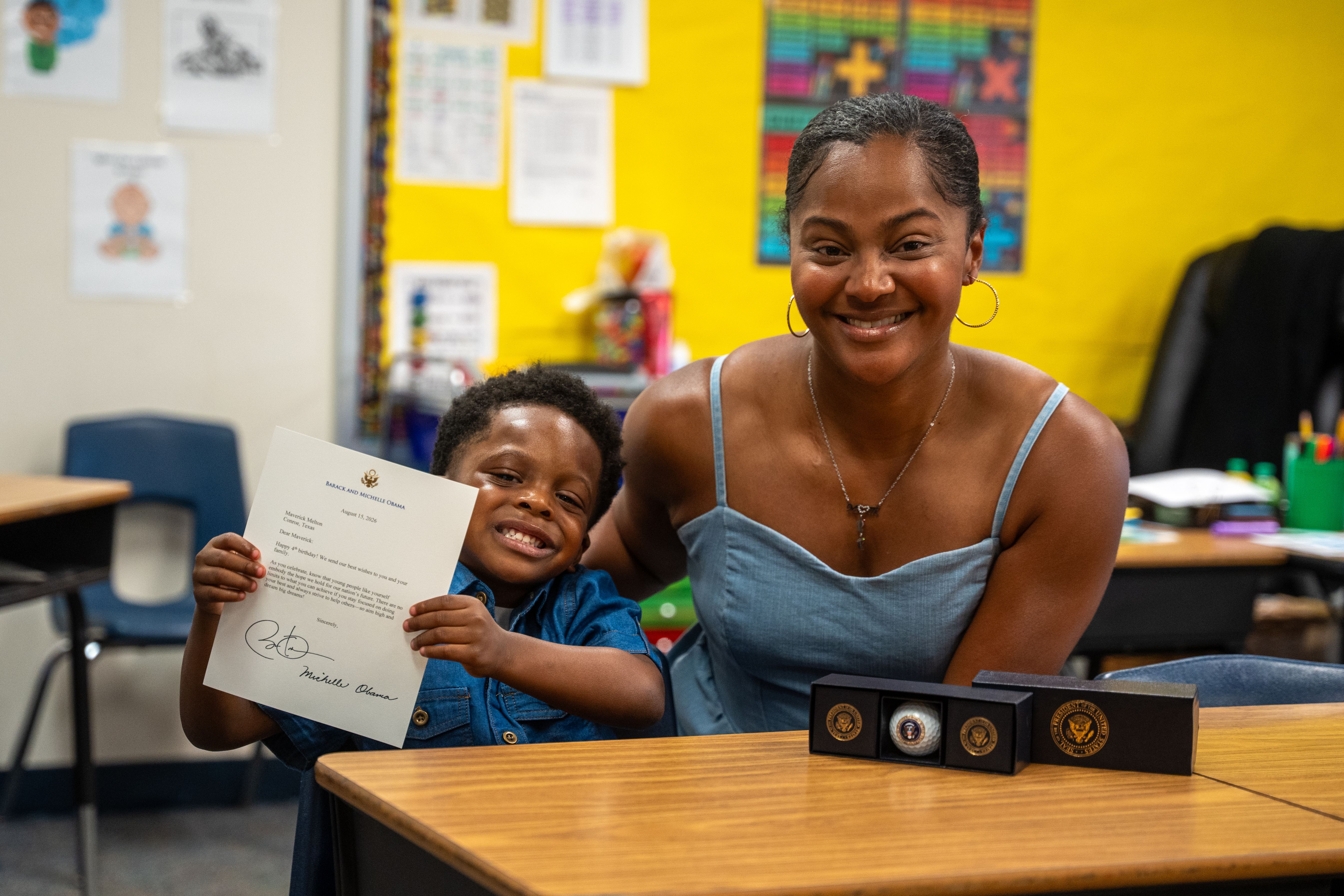 Child holding up letter smiling in classroom next to his mom.