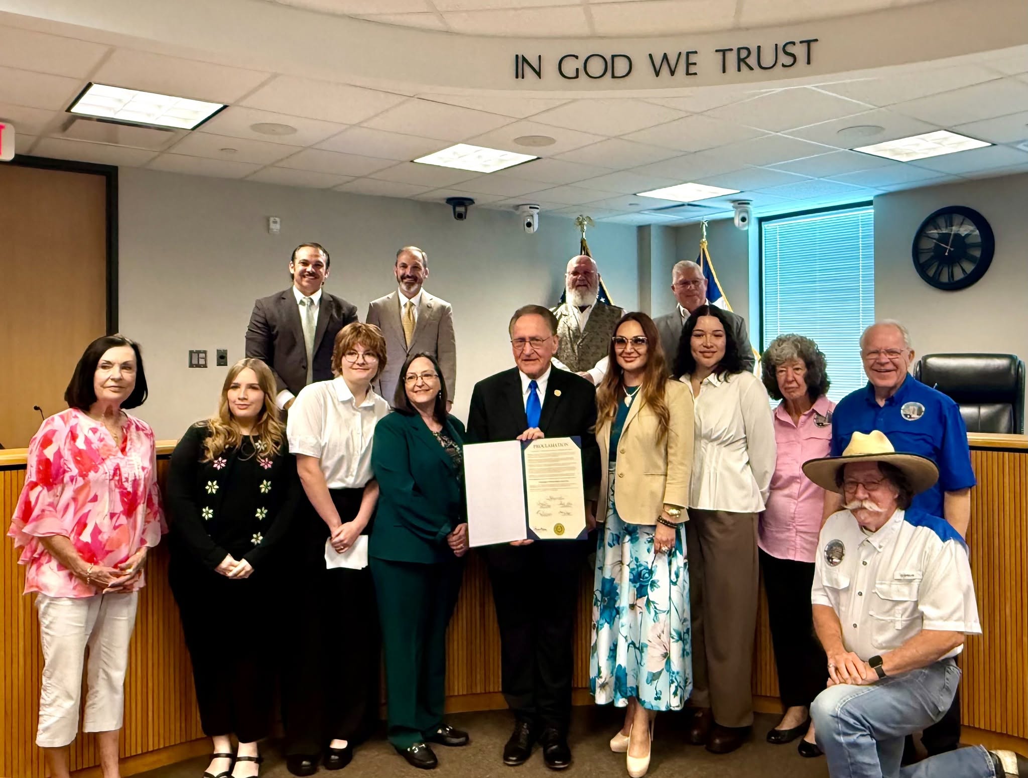 Men and women stand in city board room holding a proclomation to celebrate a business.