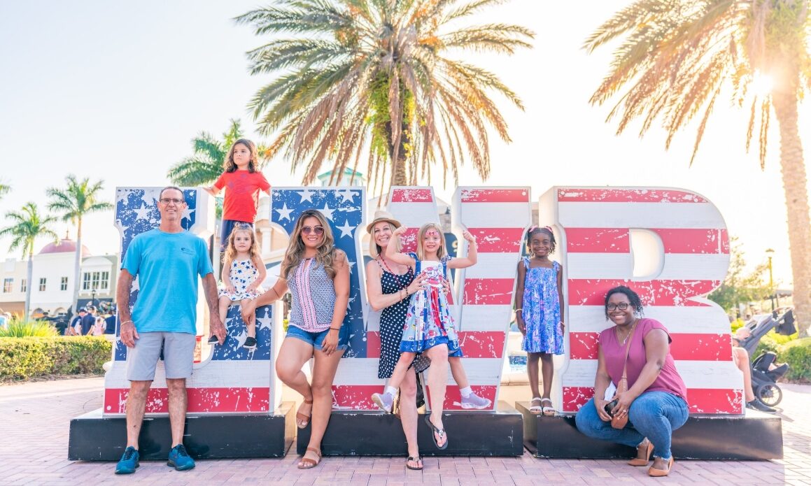 Adults and children pose beside large red, white and blue LWR letters beneath palm trees.