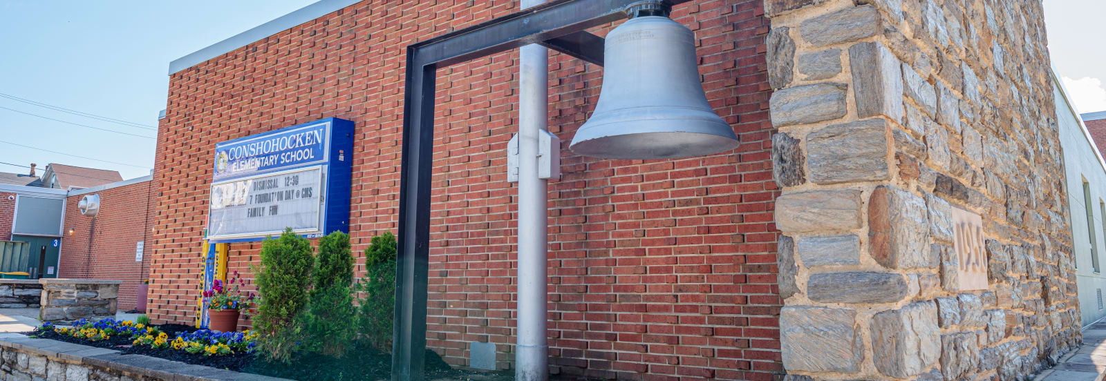 Conshohocken Elementary School’s brick-and-stone exterior, with a school sign, flower bed and bell.