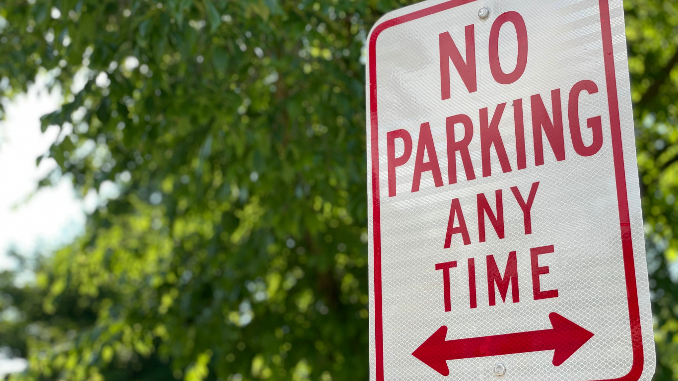 A close-up of a red-and-white “No Parking Any Time” sign with arrows pointing left and right, set against a leafy green background.