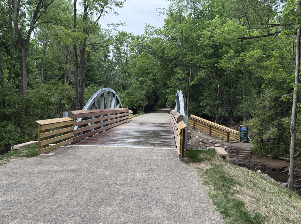 A wooden pedestrian bridge with steel arch supports crosses Paint Creek along a gravel section of the trail, surrounded by green trees.