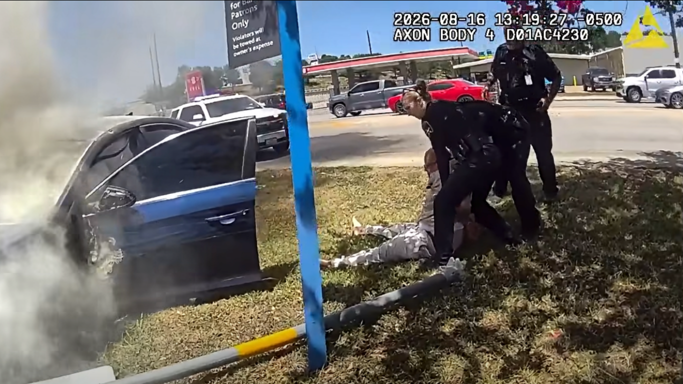 Body camera footage shows two sheriff's deputies pulling a person from the grass near a smoking, crashed sedan, with a patrol vehicle and gas station visible in the background.