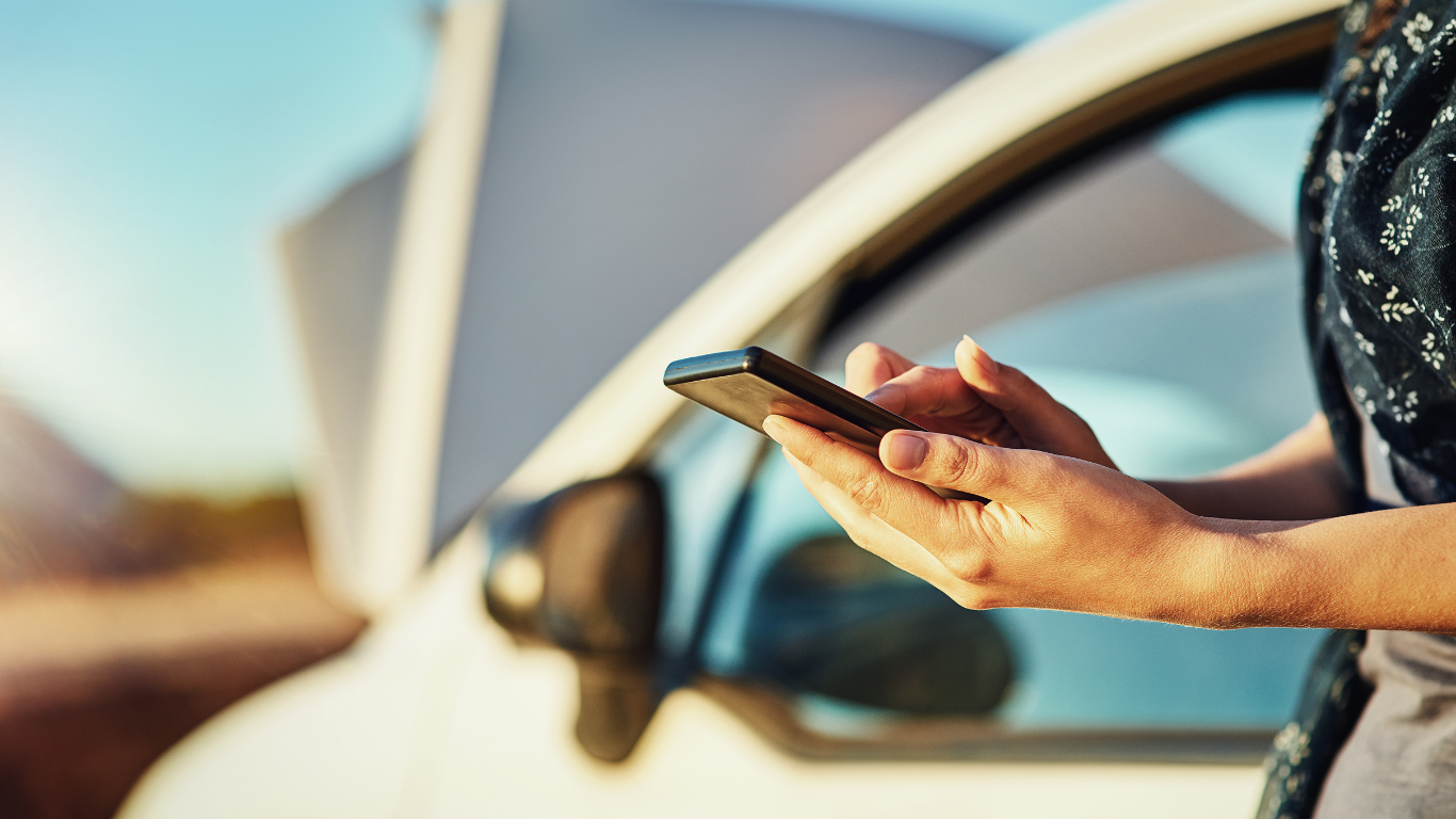 Person holding a smartphone beside an open car door, appearing to seek roadside assistance.
