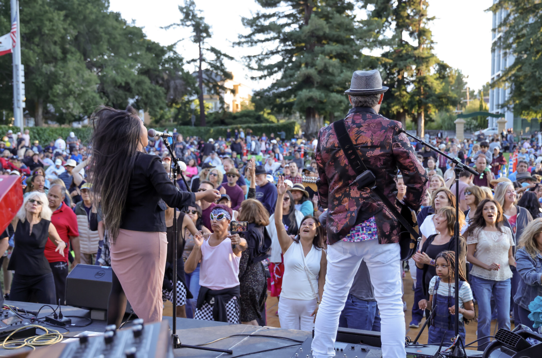 A singer and guitarist perform on an outdoor stage at Central Park in San Mateo, backs to the camera, facing a large crowd of people dancing and cheering under trees at sunset