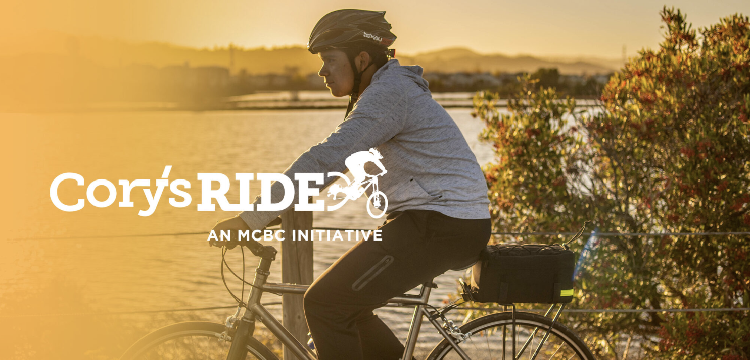 A young cyclist wearing a helmet rides a bike along a waterfront path at sunset, with the "Cory's Ride" and "An MCBC Initiative" logo overlaid on the image