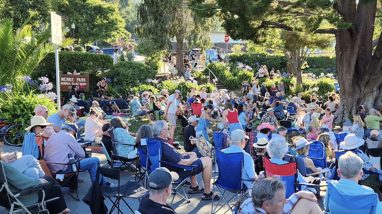 A large crowd sits in lawn chairs and on blankets under trees at an outdoor concert near the Menke Park sign in Corte Madera.
